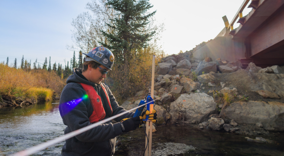Man in safety gear standing in a waist-deep rocky stream, focusing on his measuring tools.