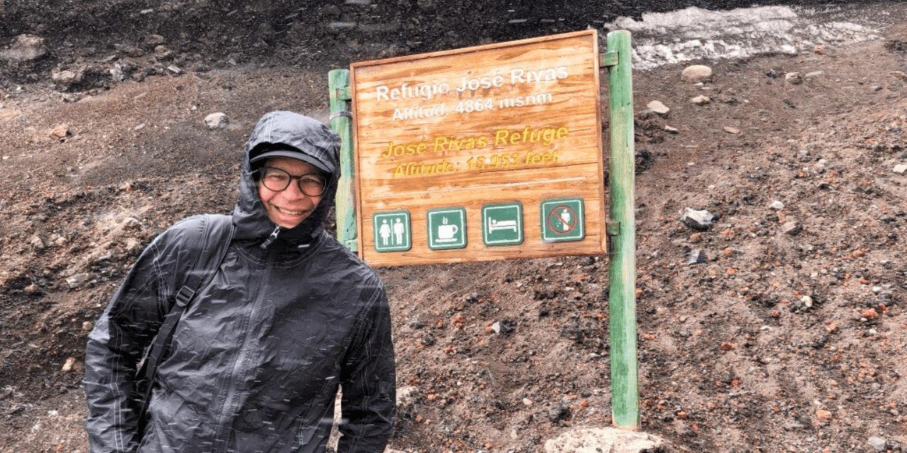 A person in a black rain jacket stands next to a wooden sign indicating "Refugio José Rivas" at a high altitude, with rocky terrain in the background.